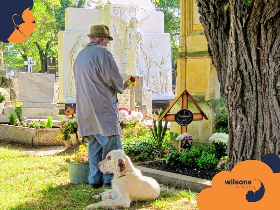 Elderly man with hat holds flowers at grave in sunny cemetery, accompanied by a white dog. Tombstones and trees in background. Wilsons Health logo.