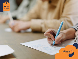 Hands writing with pens on paper at a table, blurred background. Orange and blue Wilsons Health logo visible. Calm, focused mood.
