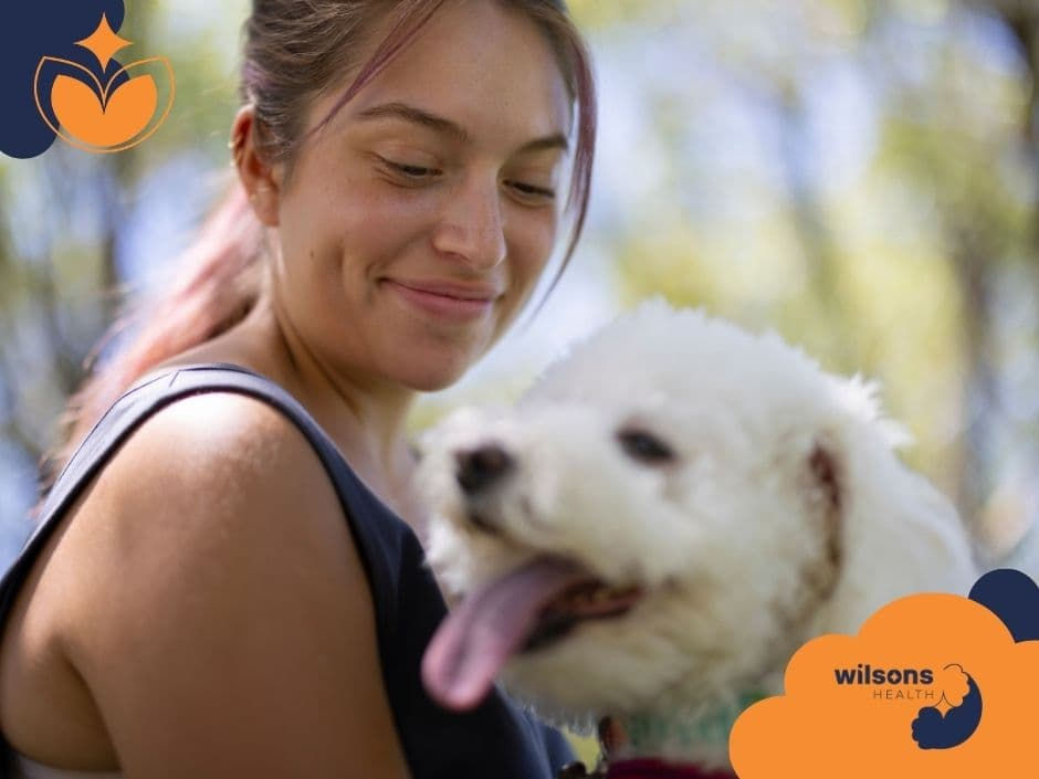 Woman smiling at a fluffy white dog outdoors. The dog appears happy with its tongue out. Tree backdrop. "Wilsons Health" logo visible.