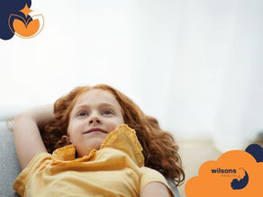 Young girl with red hair in a yellow shirt lying back and smiling. Bright room with logo "wilsons HEALTH" in the corner. Relaxed mood.