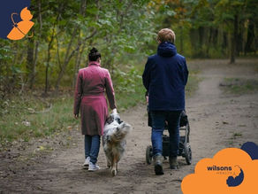 A couple walks a dog and pushes a stroller along a forest path. The woman wears a pink coat, and the man a blue jacket. Wilsons Health logo visible.