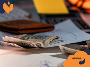 Banknotes and papers on a cluttered desk with a wallet and laptop in the background. Warm lighting, Wilsons Health logo visible.