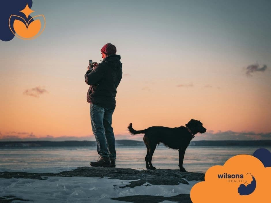 Person in winter clothing takes a photo at sunset, standing on a snow-dusted shore with a dog. Orange and navy graphics and text: "wilsons HEALTH".