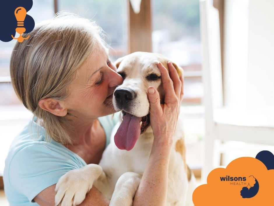 Woman lovingly embraces a dog indoors. Dog's tongue out, relaxed. Background is bright. "Wilsons Health" logo in corner.