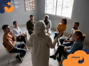 A group therapy session with diverse adults seated in a circle. A woman with gray hair leads the discussion. Bright room with "Wilsons Health" logo.
