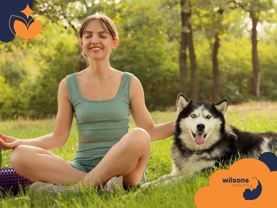 Woman in green outfit meditates beside happy husky on grass in a sunny park. "Wilsons Health" logo in corner. Relaxed and cheerful vibe.