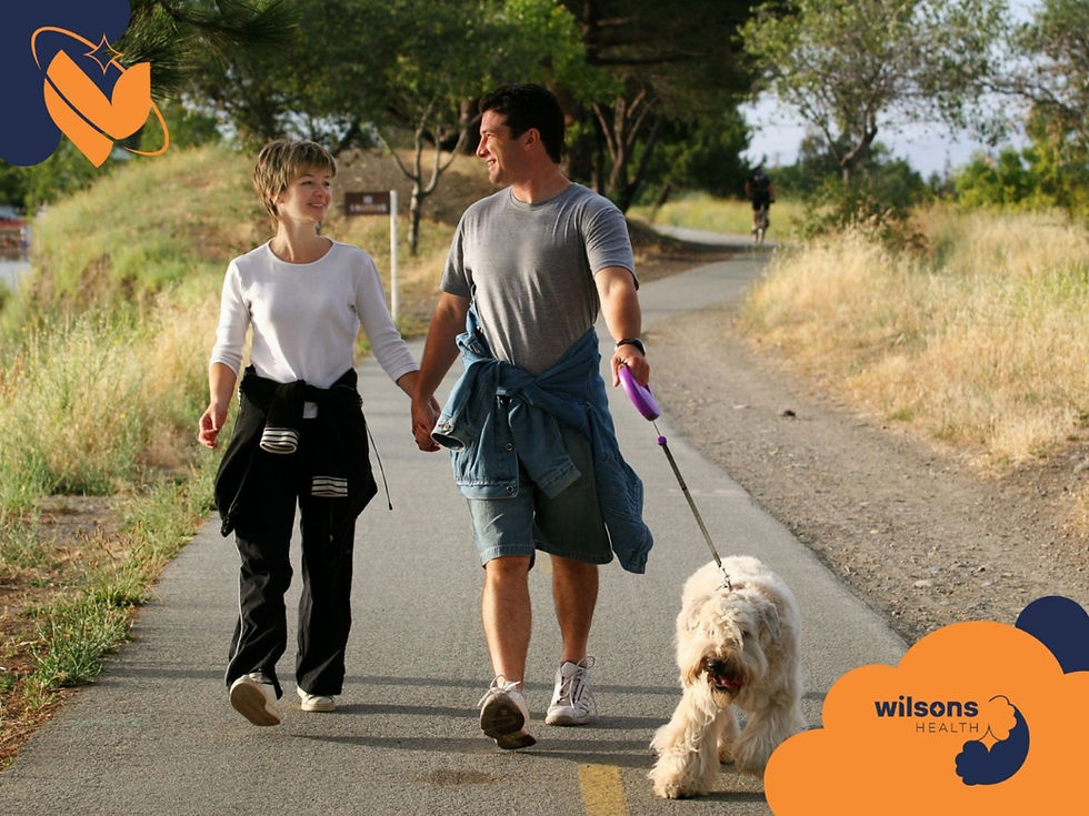 Couple holding hands, walking dog on a sunny path. Smiling, casual attire. Grass and trees in the background. Wilsons Health logo visible.