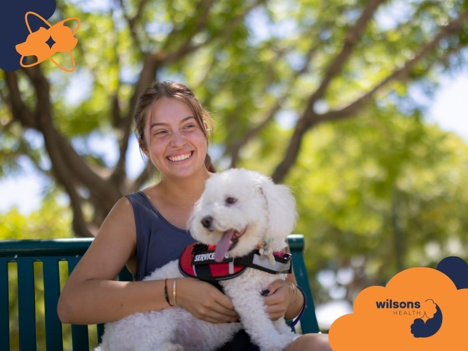 Woman smiling on a bench with a white service dog in her lap. Sunny park setting, green foliage, "wilsons HEALTH" text in corner.