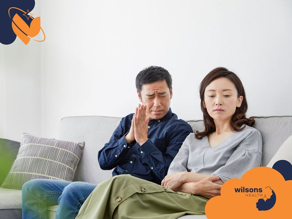 A man apologizes with clasped hands to a woman who looks away, both seated on a couch. Wilsons Health logo is visible. Mood is tense.