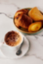A white cup of cappuccino with cocoa powder beside a plate of assorted pastries on a marble surface, creating a cozy breakfast scene.