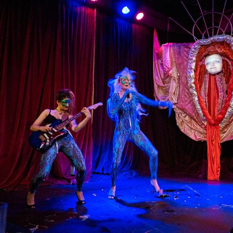 A still from the production ‘Queen C*nt’. Two performers in heels and glittery masks are performing on a dark stage: one with a guitar and one with a microphone. Behind them is a large structure with cloths and materials hanging from it, in the shape of a vulva