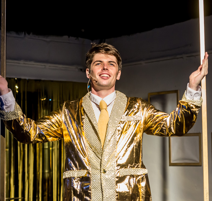 A performance still from 'The Quentin Dentin Show'. Luke Lane as Quentin Dentin is wearing a gold suit and is smiling and singing.