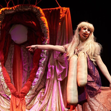 A still from the production ‘Queen C*nt’. A woman in a costume exaggerating her body features performs and points behind her to a large structure with cloths and materials hanging from it, in the shape of a vulva.