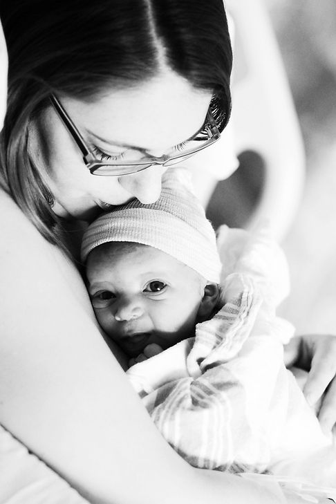A black and white image showing a side view of a mother holding her baby close to her chest. The mother, wearing glasses, is gently kissing the baby’s head. The baby, with eyes wide open and wearing a woolen hat, is nestled against her. The mother gazes lovingly at the baby as they both stand upright.