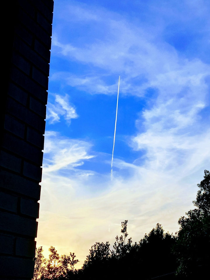 Una lunga striscia bianca, scia di un aereo, che squarcia una piccola porzione di cielo tinta di blu, tra nuvole ovattate e tenui raggi di sole.