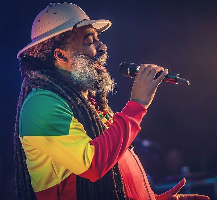 Teacha Dee in red, gold and Green rasta Jacket singing on stage