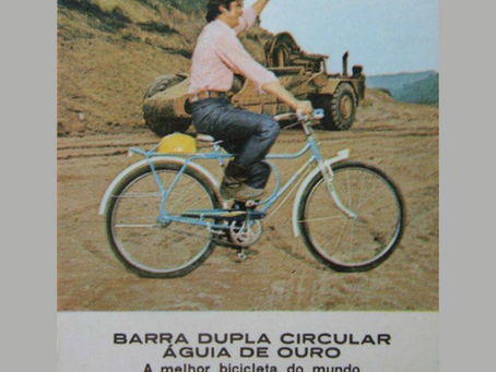 Homem sorrindo em uma bicicleta Monark azul e branca, modelo barra dupla circular 'Águia de Ouro', em um cenário de terra com máquinas de construção. Imagem vintage promocional da Bicicletas Monark SA