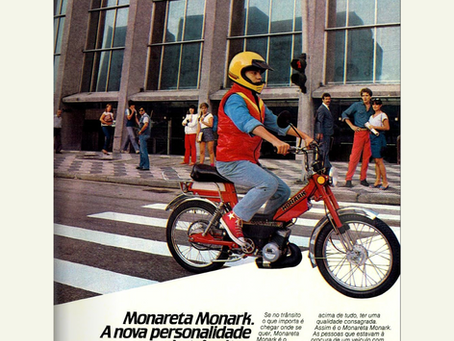 Post de tbt com uma pessoa com capacete amarelo e colete vermelho pilotando uma bicicleta Monareta Monark vermelha em uma faixa de pedestres, com um prédio moderno e pessoas ao fundo. O anúncio da Bicicletas Monark SA apresenta o slogan "Monareta Monark. A nova personalidade do trânsito." e texto publicitário sobre o veículo da Bicicletas Monark SA.