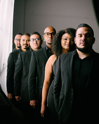 Eclectik Band, dressed in black, standing in a line, with a soft white light illuminating them from the left.