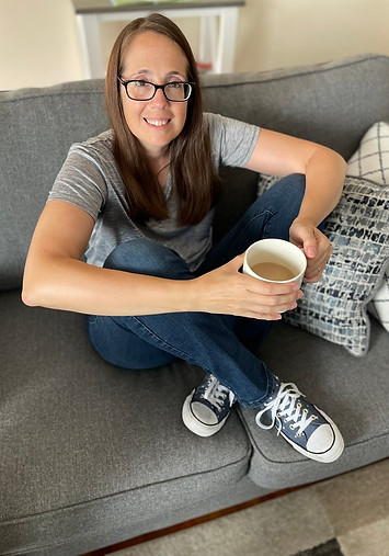 MiaMaria a woman sitting cross-legged on acouch smiling holdoing coffee in grey shirt blue jeans and converse sneakers and glasses with medium brown hair