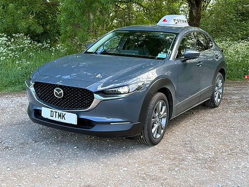 Grey Mazda learner-driver car with a DTMK roof sign, parked on a gravel road beside woodland.