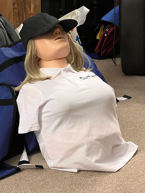 Female adult CPR training manikin with realistic chest anatomy, dressed in a white DTMK polo shirt and cap, seated upright against a chair to represent a clothed unresponsive casualty scenario used in first aid and resuscitation training.