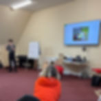 A first aid trainer stands at the front of a classroom delivering a session, with learners seated on the floor and a presentation displayed on a screen alongside training equipment.