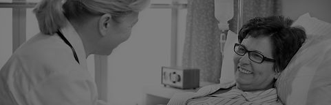 Healthcare professional speaking supportively with an older patient in a hospital bed.
