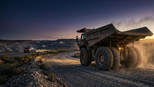 An autonomous mining haul truck on a graded haul road at blue hour, with the silhouette of a sensor-equipped autonomous passenger vehicle visible in the soft middle distance — illustrating the transfer of safety frameworks from on-road autonomous vehicles to autonomous mining