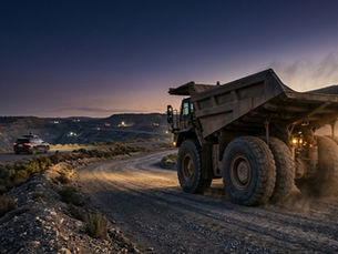 An autonomous mining haul truck on a graded haul road at blue hour, with the silhouette of a sensor-equipped autonomous passenger vehicle visible in the soft middle distance — illustrating the transfer of safety frameworks from on-road autonomous vehicles to autonomous mining