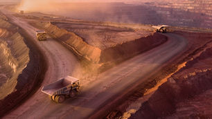 Aerial view of mining site with large trucks on winding dirt roads at sunset. Red earth tones dominate the landscape. Dust rises in the air.