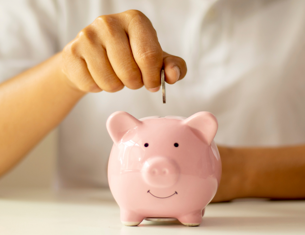 A hand places a coin into a smiling pink piggy bank on a white surface, suggesting saving or financial planning.