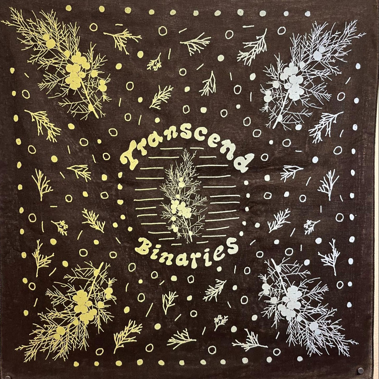 Black handkerchief or “hanky” with light print. The words transcend binaries encircle a representation of a plant with berries that appears elsewhere on the hanky with circles and dashes surrounding it.