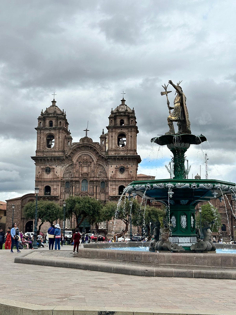 People walk near a fountain and historic church under cloudy skies. The fountain features a statue and is surrounded by trees and architecture.