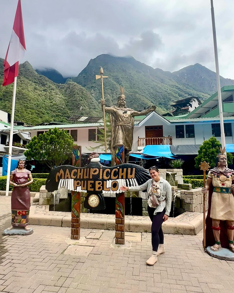 Person posing by 'Machupicchu Pueblo' sign with Inca statues, surrounded by mountains and buildings. Overcast sky, Peruvian flag visible.