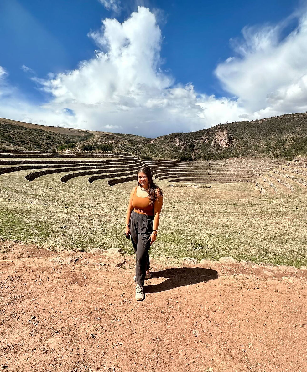 A woman in orange top walks on reddish dirt path, with ancient terraced landscape and cloudy blue sky in the background. Mood is cheerful.