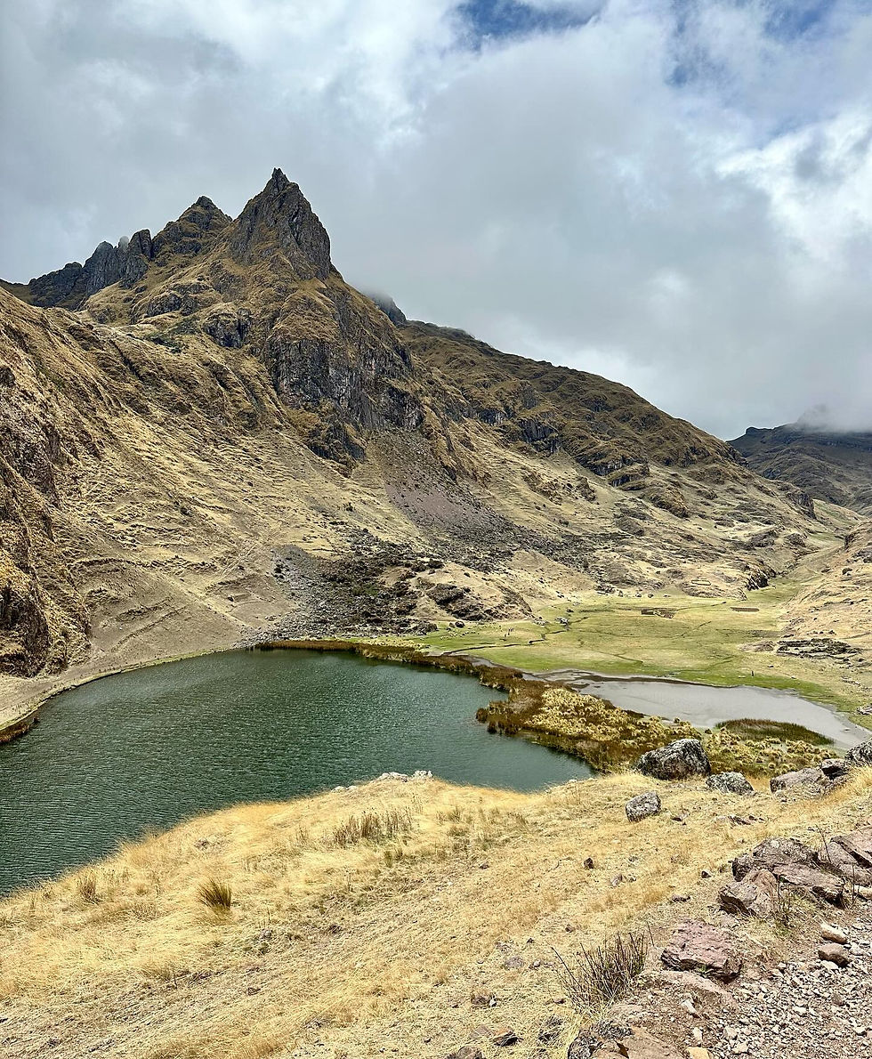 Mountainous landscape with a rocky peak, grassy hills, and a small lake. Cloudy sky sets a serene mood. Earthy tones dominate the scene.
