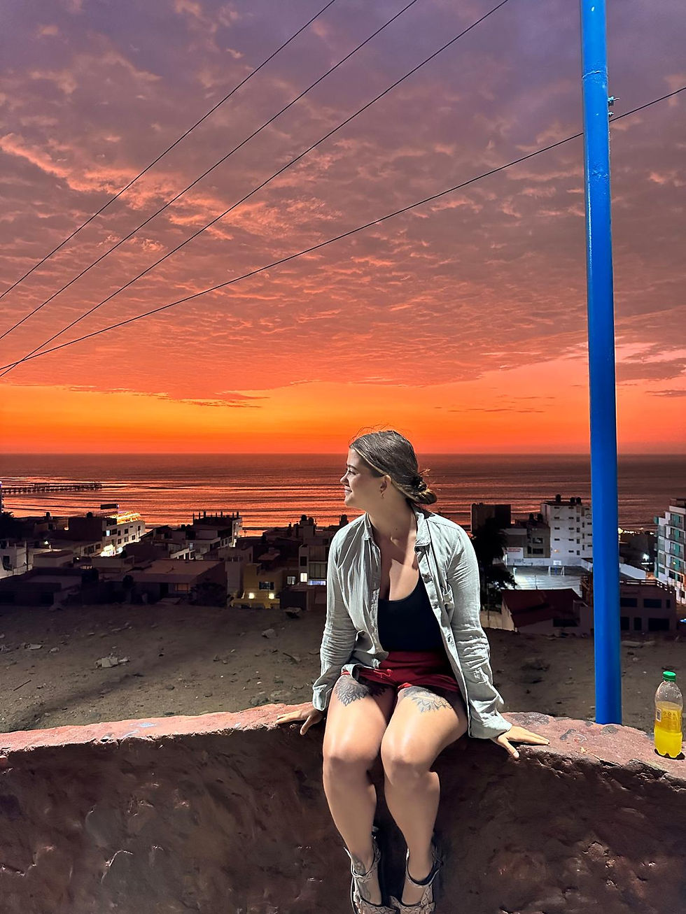 A woman sits on a ledge, gazing at a vivid orange sunset over a coastal town. A blue pole is nearby, with a drink bottle on the wall.