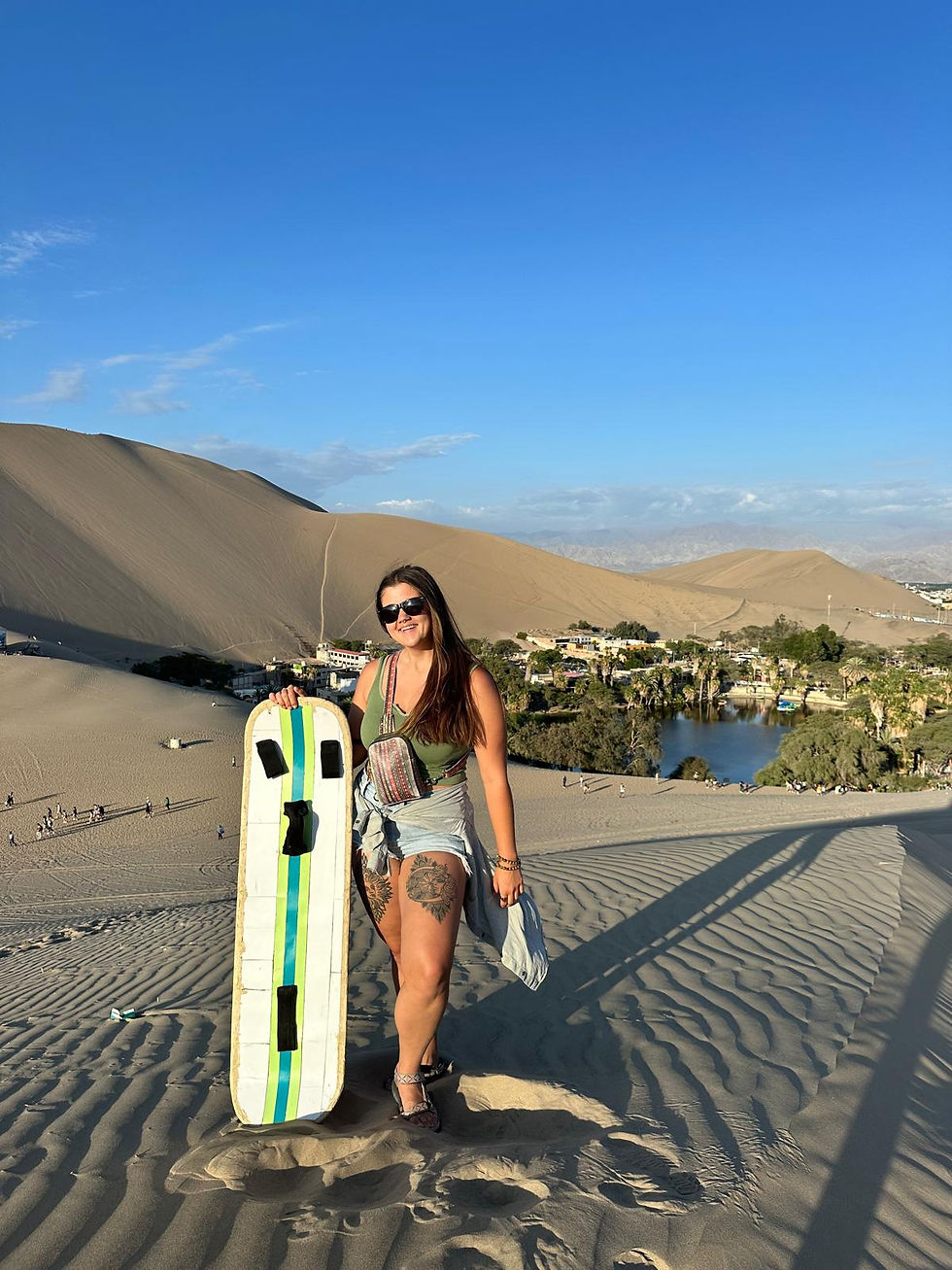 Woman in a desert holding a sandboard, wearing sunglasses. Sandy dunes and an oasis are in the background under a clear blue sky.