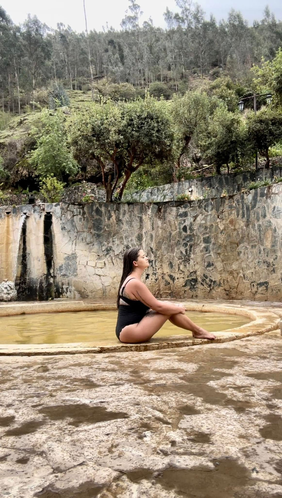 Woman in a black swimsuit sits by a stone pool with a waterfall. Green forest in the background. Calm and serene mood.