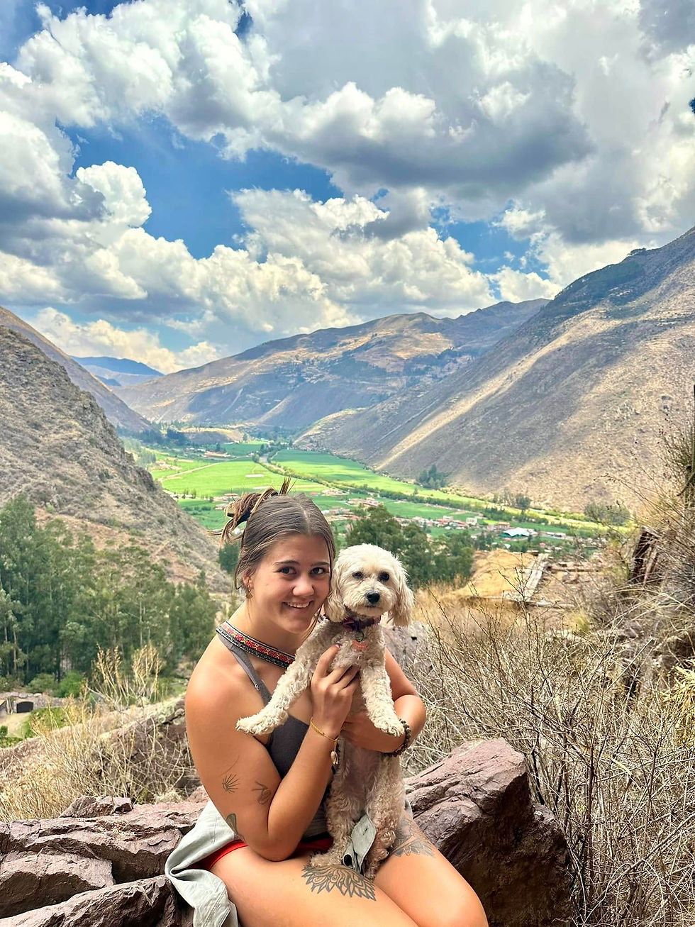 Woman smiling while holding a small dog, sitting on a rock with a mountainous landscape in the background. Cloudy sky, lush green valley.