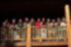 Eleven women smiling on a wooden balcony, wearing casual clothes. Bright sunlight creates a warm atmosphere against the dark background.