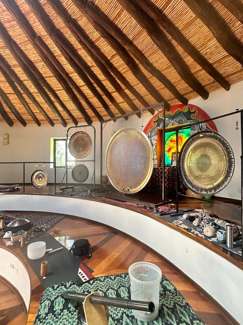 Indoor setting with hanging gongs, a colorful mural, and a wooden ceiling. Musical instruments are scattered on a patterned mat. Quiet ambiance.
