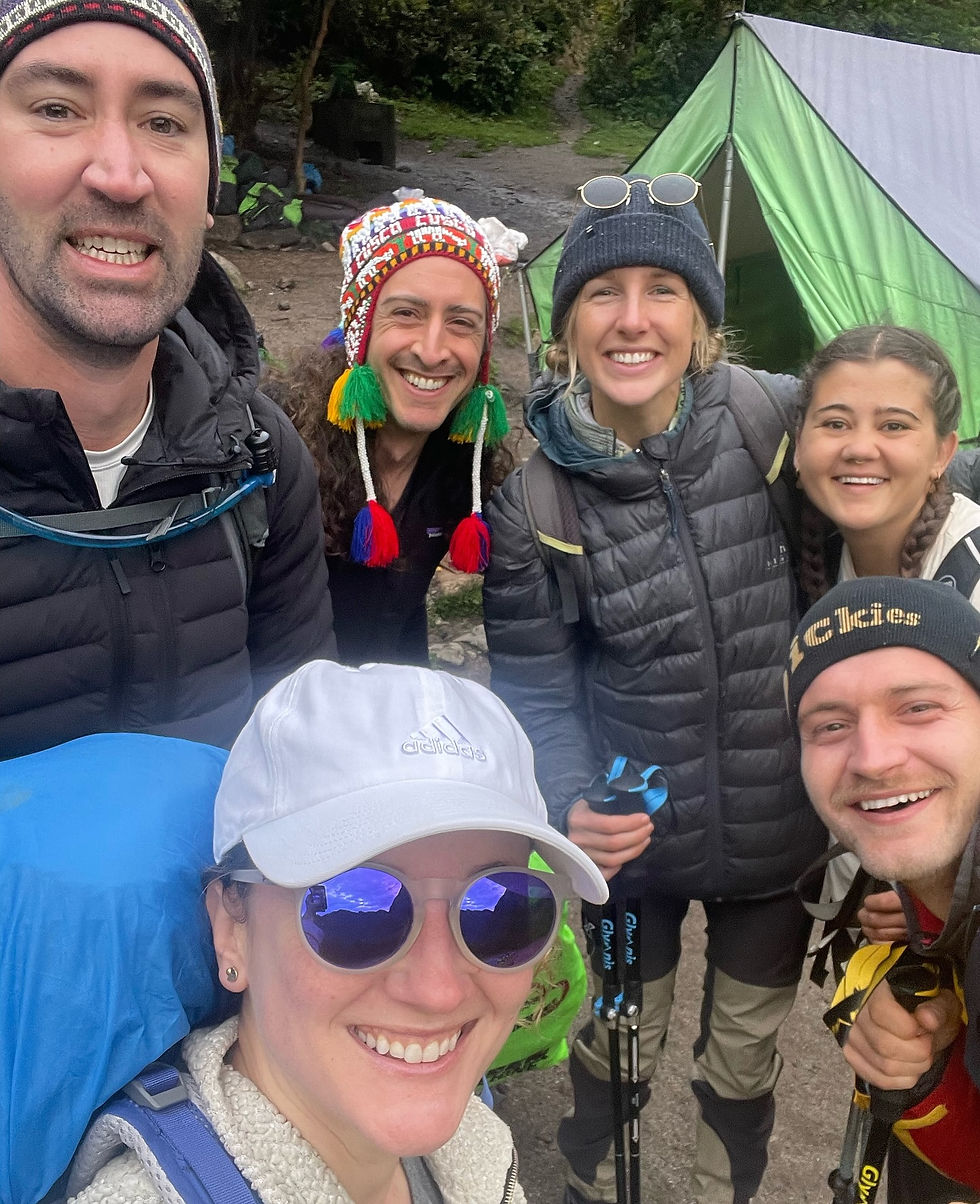 Group of six smiling hikers in winter gear pose by a green tent in a forest. One wears a colorful hat. Sky reflected in sunglasses.