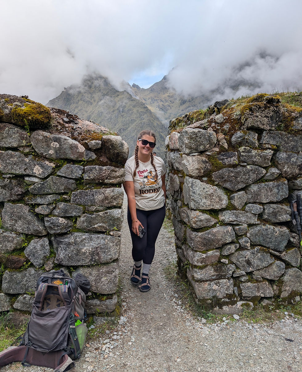 Woman in sunglasses stands between stone walls, smiling. Backpack on ground. Cloudy mountains in background. Casual, adventurous mood.