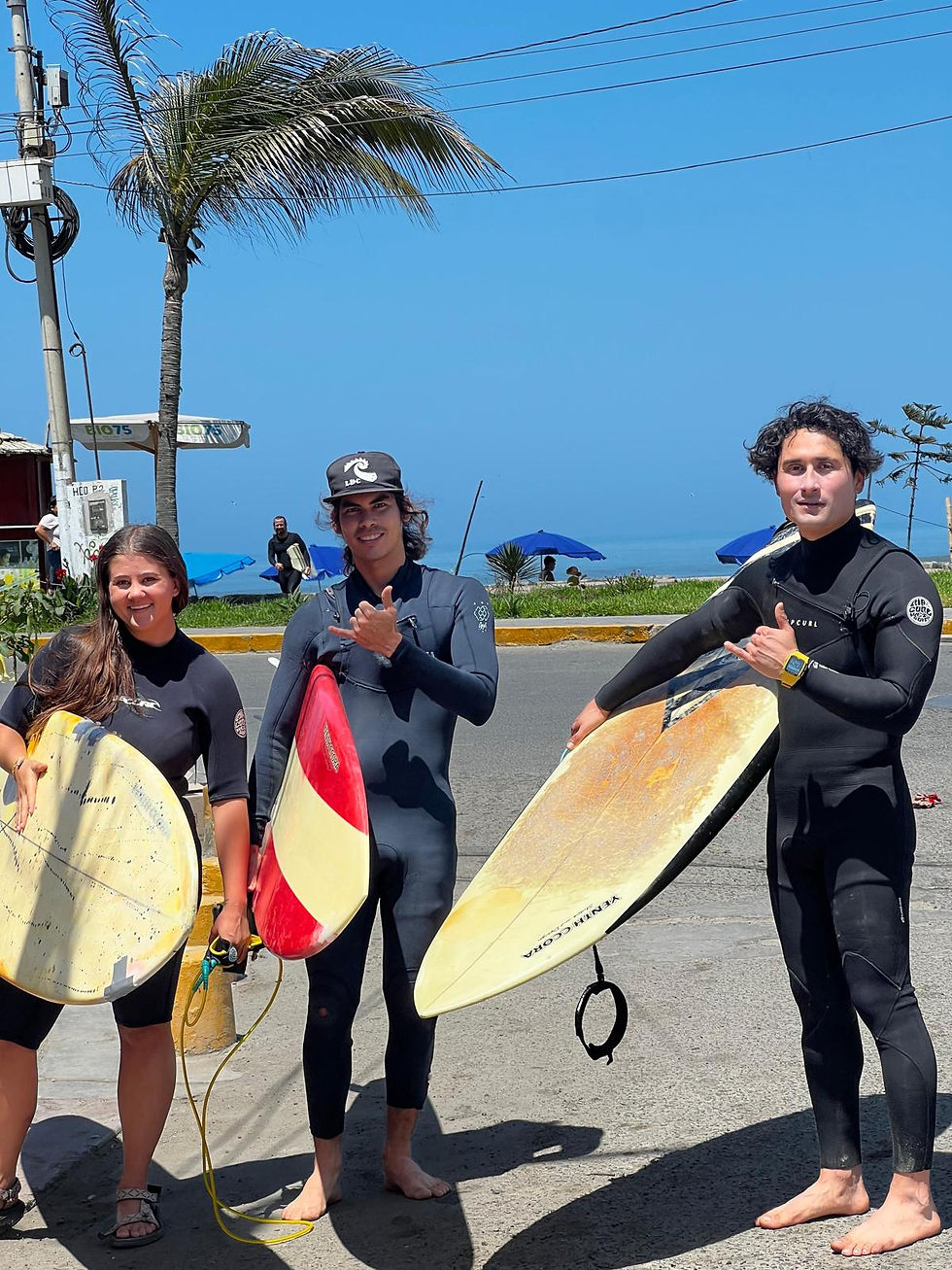 Three surfers holding boards pose with shakas by the beach, under a palm tree. Clear blue sky and ocean umbrellas in the background.