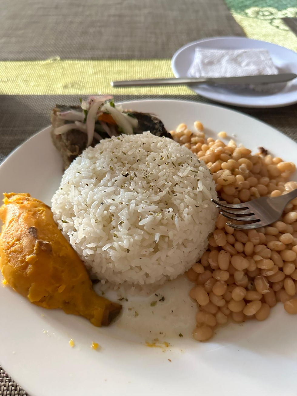 A plate of rice, beans, baked plantain, fish with onions. Cutlery on a napkin in the background on a woven placemat, daylight setting.