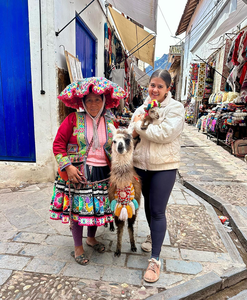 Two women with a llama and a lamb on a colorful street. One wears traditional attire, the other in white. Vibrant textiles in the background.