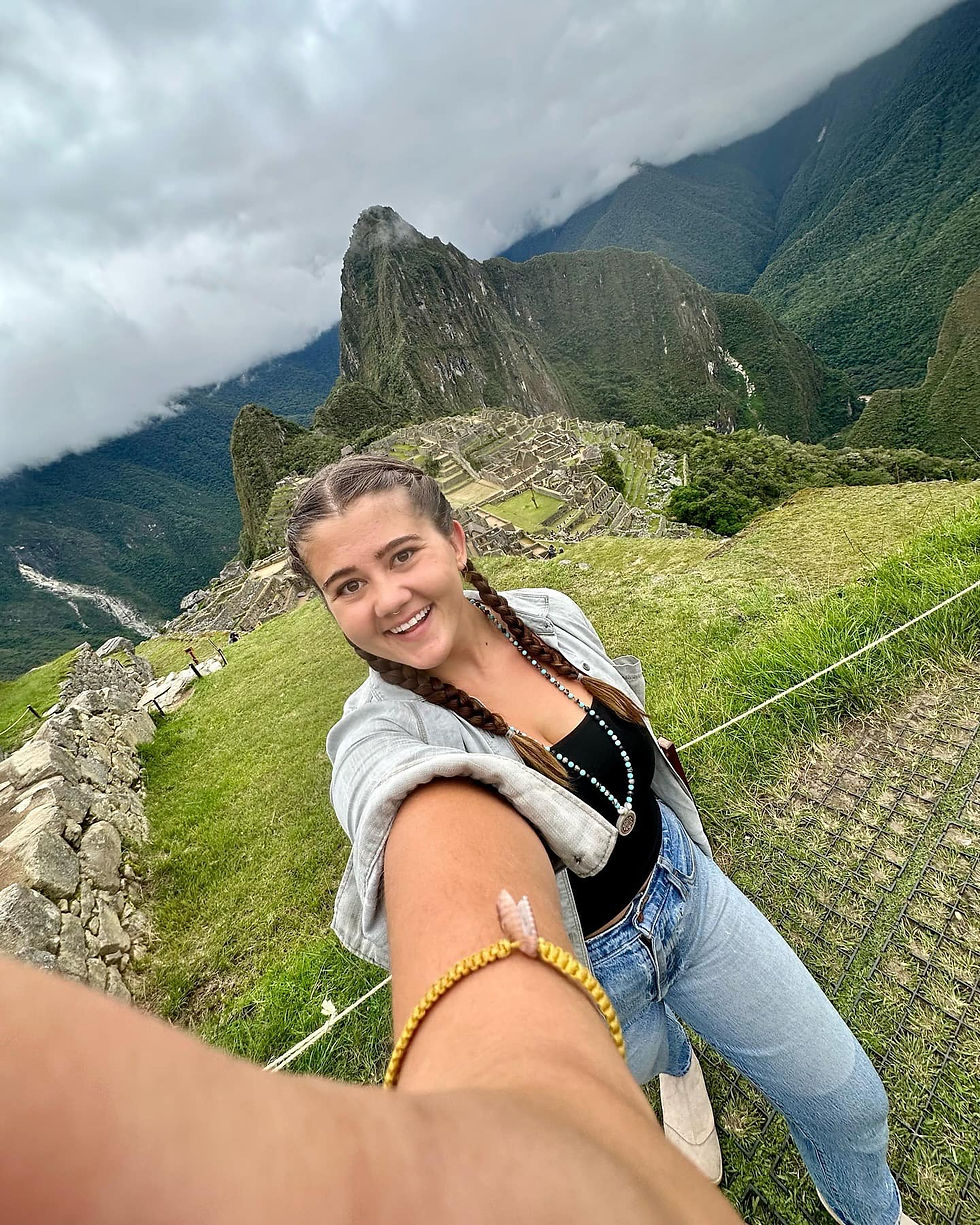 Smiling woman taking a selfie at Machu Picchu. Overcast sky, lush green mountains in the background. Relaxed, adventurous mood.