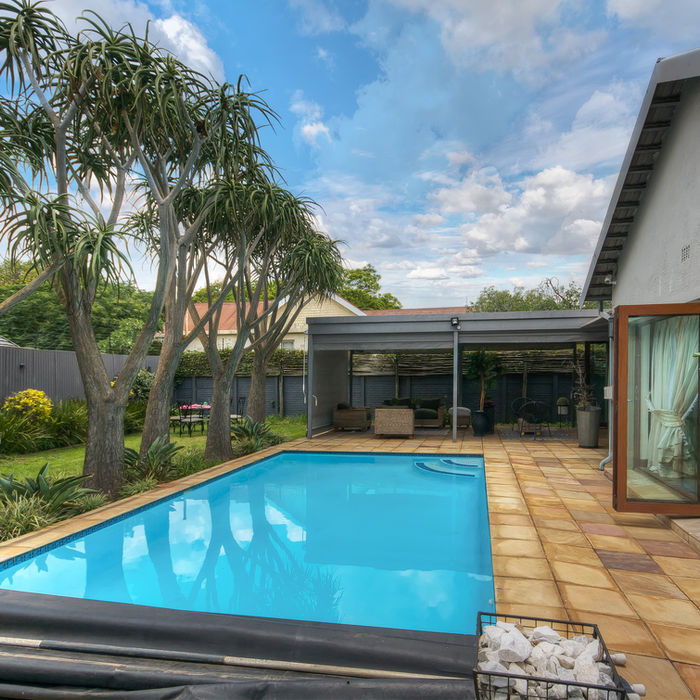 Blue pool with trees on the side, with a white house and an outside lounge area